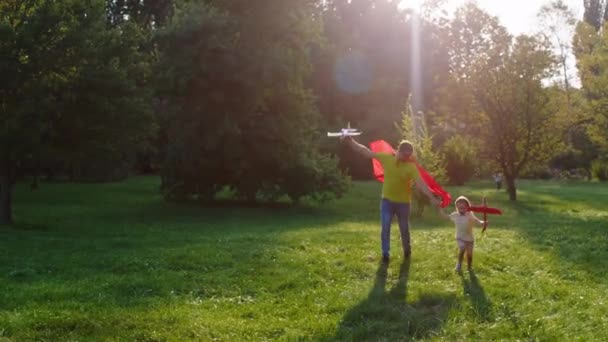 Feliz y emocionado papá con su pequeño hijo caminando a través de la hierba con un padre avión rojo con traje de superhéroe — Vídeos de Stock