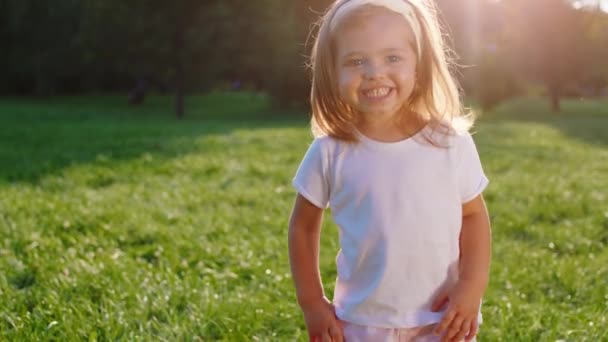 Stort leende en söt och vacker liten flicka framför kameran i parken hon tittar rakt in i kameran — Stockvideo
