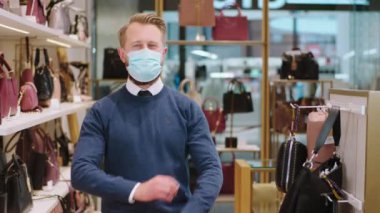 Sarışın, yakışıklı bir kuaför kameranın önünde el ele tutuşup Coronavirus salgınında maske takıyor ve aksesuar dükkanının ortasında duruyor.