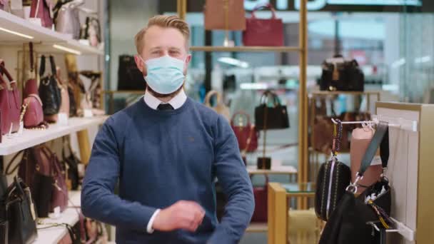 Snygg blond hårförsäljare poserar framför kameran korsar händerna och bär mask i pandemin av coronavirus han står i mitten av tillbehör butik — Stockvideo