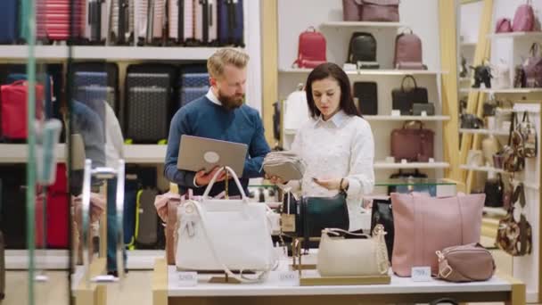 Devant la caméra superviseur charismatique homme utilisant un ordinateur portable pour vérifier le prix des sacs avec la vendeuse dans la boutique d'accessoires — Video