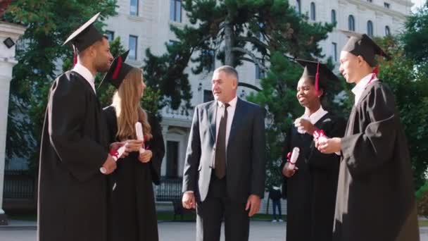 Szczęśliwy profesor koledżu pogratulował swoim studentom ukończenia szkoły w ogrodzie koledżu wszyscy razem są podekscytowani trzymając ich dyplomy — Wideo stockowe