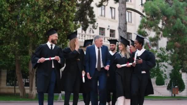 Ευτυχισμένοι και ενθουσιασμένοι απόφοιτοι φοιτητές που περπατούν μπροστά στην κάμερα μαζί με τον διευθυντή του κολεγίου μετά την αποφοίτηση συζητούν όλοι μαζί — Αρχείο Βίντεο
