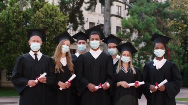 Вне группы выпускников колледжа мультирасовые студенты после окончания школы позируют перед камерой в группе в защитной маске они находятся в пандемии COVID-19 — стоковое видео