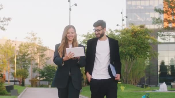 Marcher devant la caméra deux étudiants charismatiques dame et gars discutant tenant une tablette électronique et d'analyser quelque chose de fond bâtiment d'université moderne — Video