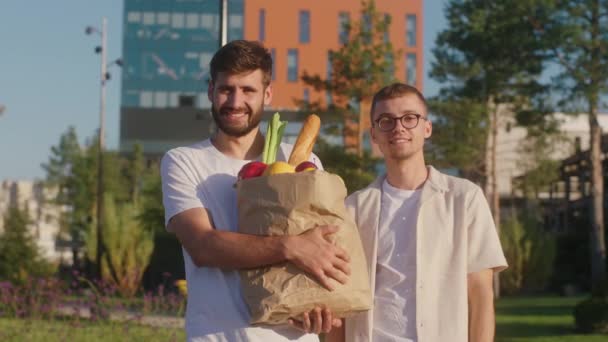 Modern bir kasabada sokağın ortasında kameranın önünde poz veren iki karizmatik adam. Biri yiyecek dolu bir çanta taşıyor. — Stok video
