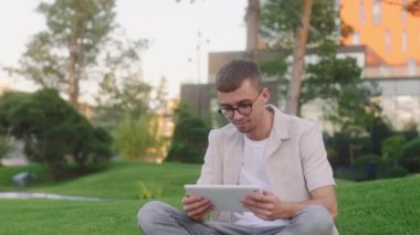 Tablet kullanan yakışıklı genç bir öğrenci üniversite parkında çimlerin üzerinde bir nesne için proje yapıyor.