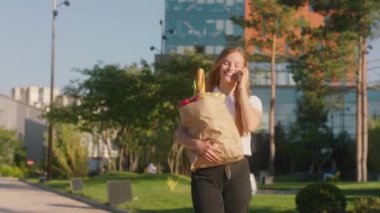 Kameranın önünde yürüyen sarışın kadın elinde sebze ve meyve dolu bir çantayla sokakta yürüyor telefonda konuşuyor ve güneşin tadını çıkarıyor. 4k