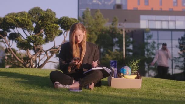 草の上にダウン公園の真ん中に座って、誰かを呼び出すために開始大きな現代的なビジネスセンター — ストック動画