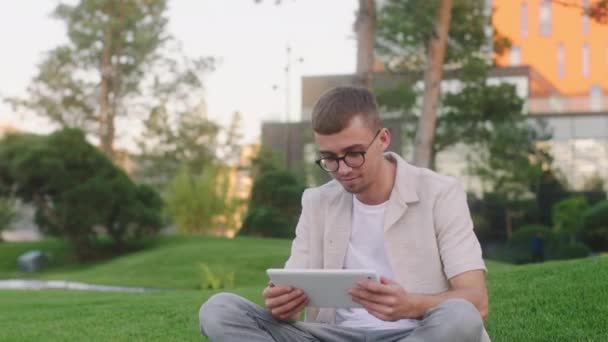 Guapo chico joven estudiante usando una tableta para hacer un proyecto de algún objeto en el parque de la universidad en la hierba — Vídeos de Stock