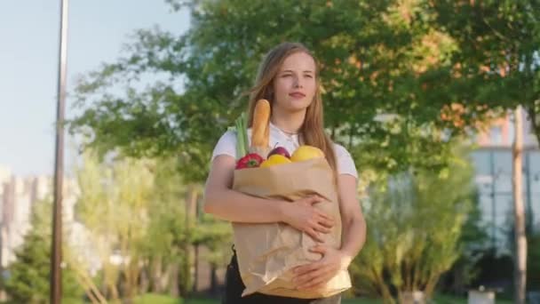 An einem sonnigen Tag vor der Kamera geht eine gut aussehende Dame nach dem Einkaufen mit einer Öko-Einkaufstasche voller Gemüse und Obst die Straße entlang — Stockvideo