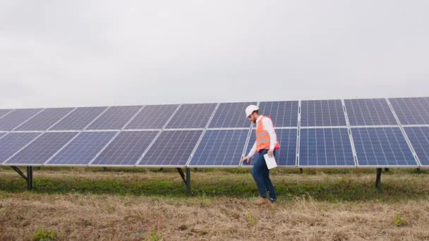 太陽光発電所の農場で安全ヘルメットとカリスマ生態技術者彼は太陽電池の清浄度をチェックし、太陽電池パネルを歩く — ストック動画