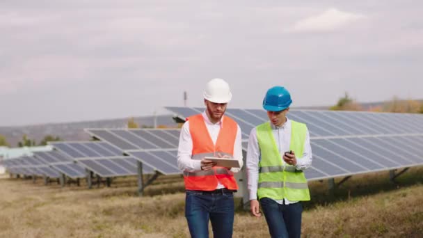 Concepto de innovación eléctrica dos trabajadores ecológicos multirraciales con equipo de seguridad caminando frente a la cámara a través de la central eléctrica de paneles solares y discutiendo algo — Vídeo de stock