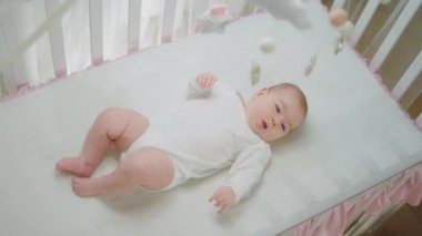 Bebek ve çocukluk kavramı. Güzel kız bebek karyolasına uzanıp, sevimli bacaklarıyla oynayan ve hareket eden bebek oyuncaklarına bakıyor. 4k