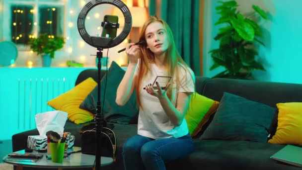 Bella giovane donna bellezza blogger facendo un trucco passo dopo passo e registrando sul suo smartphone utilizzando un anello leggero lei seduta sul divano a casa — Video Stock