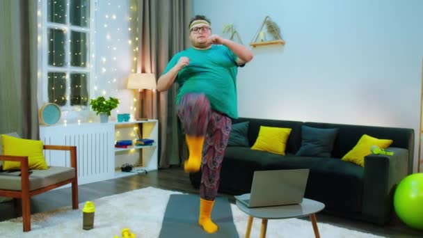 An obese man is on a blue workout mat in a living room, he is doing front kicks and looking at his laptop as he is getting help from his online trainer, he seems happy with himself — Stock Video