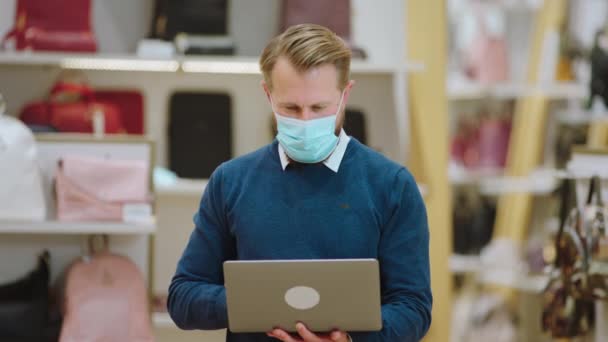 Karizmatikus eladó a kiegészítők boltjában, védőmaszkot visel, és a laptopján dolgozik a kamera előtt a koronavírus járvány idején. — Stock videók