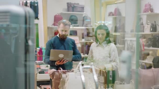 Prendre la vidéo de la vitrine d'un magasin de deux travailleurs utilisant un ordinateur portable pour faire quelques changements dans le prix discuter ensemble une dame et un homme — Video