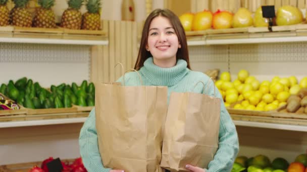 Senhora cliente feliz com eco sacos de pé na frente da câmera na loja de legumes eco segurando a sacos cheios de vegetais orgânicos e frutas — Vídeo de Stock