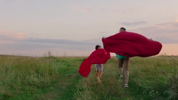 Gut aussehender Mann und sein charismatischer Sohn rennen bei Sonnenuntergang in Superheldenanzügen durch die grüne Wiese mitten in der Natur — Stockvideo