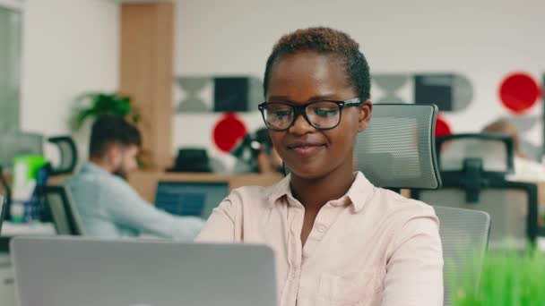 Eine junge intelligente und attraktive Afrikanerin sitzt an ihrem Schreibtisch und strahlt, als sie ihre Arme verschränkt und in die Kamera blickt. — Stockvideo