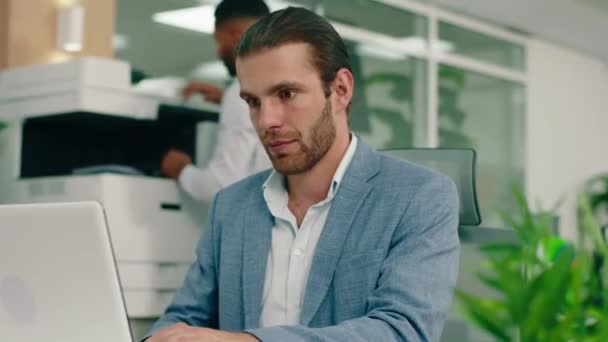 Un homme blanc attrayant avec les cheveux attachés porte un costume bleu et fait du travail sur l'ordinateur portable dans un grand bureau — Video