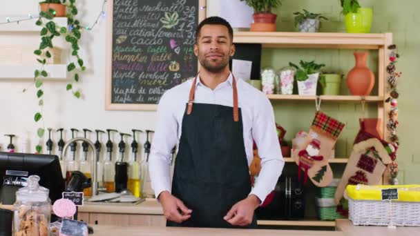 Voor de camera charismatische zwarte man barista poseren voor de camera in de coffeeshop hij kruisen handen en glimlachen een beetje. Neergeschoten op ARRI Alexa Mini. — Stockvideo