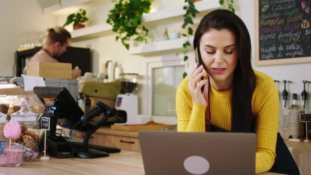 Charismatická s velkým úsměvem žena barista mluvit na smartphone se zákazníkem, že vyzvednout objednávku a typ na notebooku v útulné kavárně — Stock video