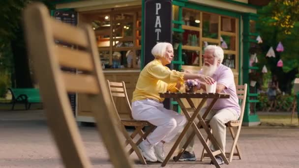 Pěkná stará žena a její manžel starý muž vzít snídani v parku v kavárně pití kávy a diskutovat — Stock video