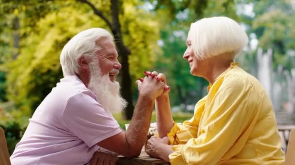 O femeie destul de bătrână și soțul ei zâmbind mare la cafeneaua din mijlocul parcului ținându-se de mână și uitându-se cu pasiune unul la celălalt. — Videoclip de stoc