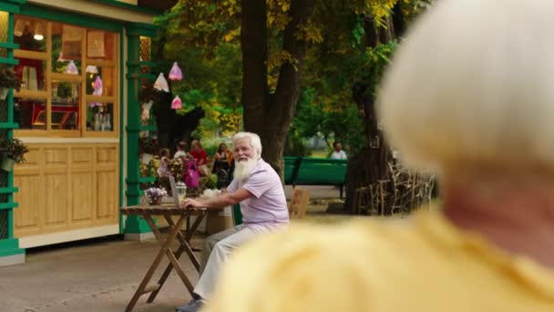 Im Café mitten im Park trifft ein romantischer alter Mann auf eine schöne Frau, während er auf dem Stuhl sitzt und ihr Luftküsse schickt. 4k — Stockvideo