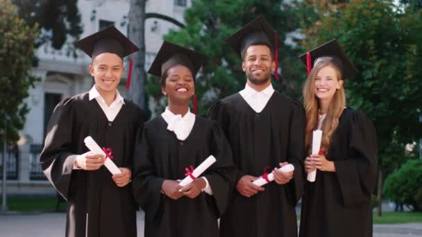 Pozowanie przed kamerą grupy absolwentów w ogrodzie studenckim trzymają swoje dyplomy i nosić czapki dyplomowe są bardzo podekscytowani wszyscy z dużym uśmiechem — Wideo stockowe