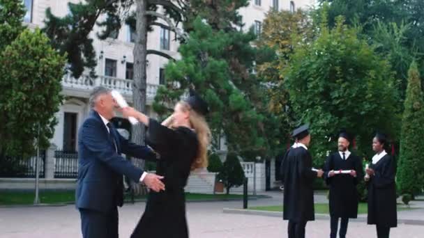 Dzień ukończenia szkoły w ogrodzie koledżu szczęśliwa pani absolwentka biegnie do ojca z dyplomem przytulając się i zdejmuje czapkę ukończenia szkoły — Wideo stockowe