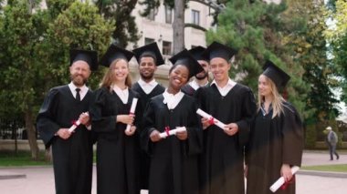 Üniversite parkındaki bir grup çok ırklı öğrenci ellerinde diplomalarla kameranın önünde büyük bir gülümsemeyle poz veriyorlar. Mezuniyet şapkası takıyorlar.