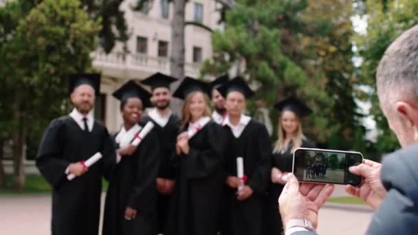 대학 공원에 있는 신나 고행복 한 졸업생들의 무리가 스마트폰 앞에 포즈를 취하고 있습니다. 학부모중 한 명 이 졸업 후 기억을 위해 사진을 찍고 있습니다. — 비디오