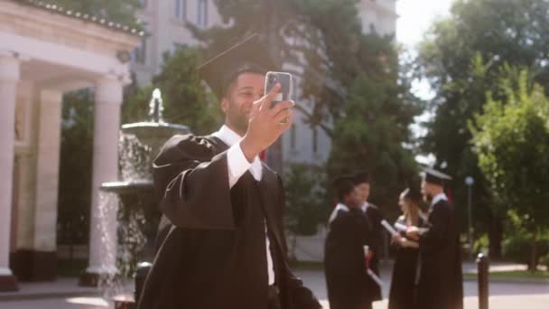 Nel giorno di laurea giardino universitario nero ragazzo alla ricerca laureato parla con qualcuno sullo smartphone mostrando sulla fotocamera il suo diploma e altri suoi colleghi — Video Stock