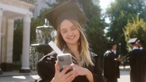 Χαμογελώντας μεγάλο χαριτωμένο φοιτητής κυρία πάρει το smartphone που κάνει ένα βίντεο για τους γονείς της μετά την αποφοίτησή της στον κήπο κολέγιο φόντο άλλη ομάδα πολυφυλετικών φοιτητών συζητούν όλοι μαζί — Αρχείο Βίντεο