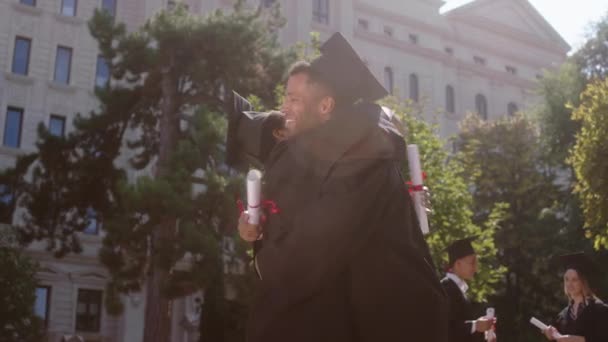 No jardim da faculdade graduados dois estudantes preto olhando jovem e mulher feliz abraçando uns aos outros e parabenizou eles discutindo enquanto segurando os diplomas vestindo bonés de formatura e ternos — Vídeo de Stock