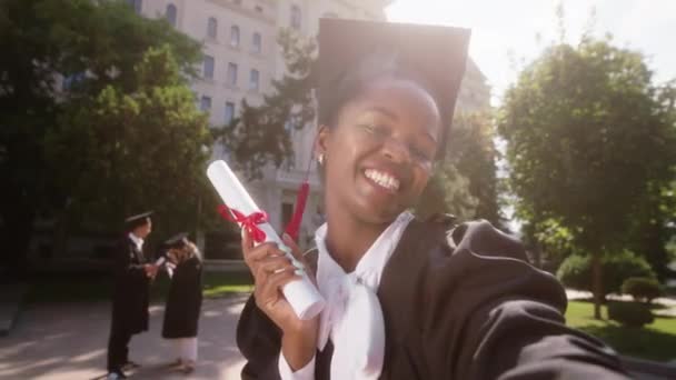 Boa aparência estudante senhora negra após a formatura no jardim da faculdade usando a câmera fazer um vídeo selfie para memórias ela mostrando o diploma muito animado sorrindo grande. Tiro em ARRI Alexa Mini — Vídeo de Stock