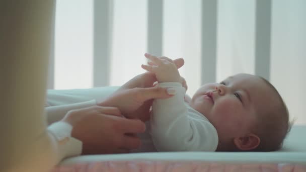 В спальне крупным планом перед камерой удивительная хорошенькая малышка обнимает мамин палец, пока малыш лежит на детской кроватке — стоковое видео