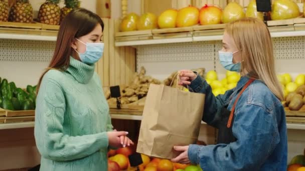 野菜屋の若い顧客と話し合う保護マスクを持つ幸せな農家の女性熟した新鮮な野菜や果物でいっぱいのエコバッグを与える — ストック動画
