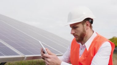Kameranın önünde, yeni modern güneş enerjisi ekoloji mühendisi dijital tablet kullanarak güvenlik kaskı takan kablosuz yondan fotovoltaik pilleri kontrol ediyor.