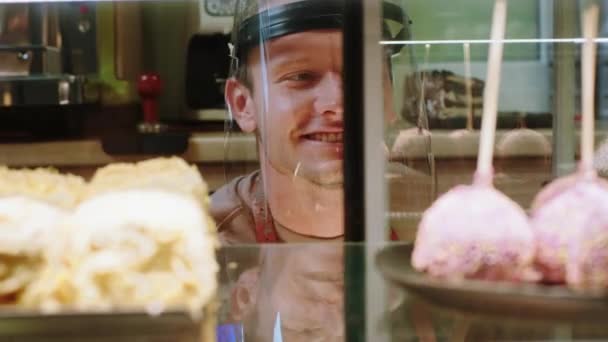 Перед камерой в пекарне кафе крупным планом улыбается симпатичный продавец с защитной маской в пандемии коронавируса аккуратно расположить свежие и вкусные десерты из витрины холодильника — стоковое видео