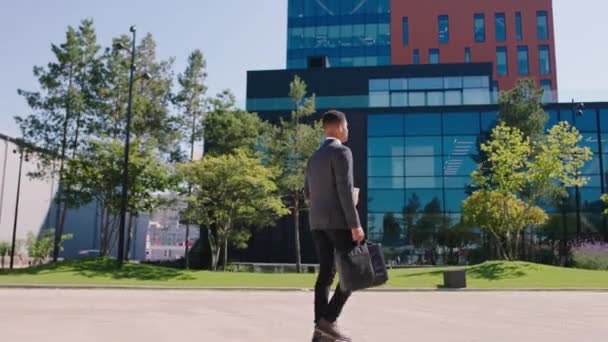 Good looking black guy office manager walking to work through the business centre park big building ahead man holding some coffee and bag. Shot on ARRI Alexa Mini. — Stockvideo