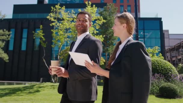 Dans une journée ensoleillée parfaite charismatique dame employée de bureau et son collègue homme noir marchant ensemble dans la rue à l'heure de pause discuter ensemble à côté du centre d'affaires moderne sur le fond — Video