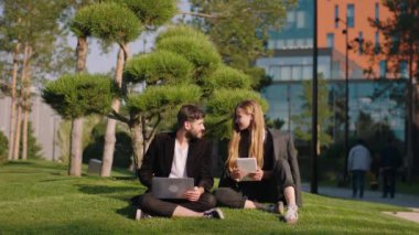 Mükemmel güneşli bir günde modern bir parkta çimlerde dinlenen güzel bir çift birlikte bir şeyler izlemek için dizüstü bilgisayar ve tablet kullanıyorlar.