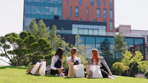 Šťastná a vzrušená skupina krásných dam po dobrém nákupním dni se posaďte na trávu uprostřed moderní zahrady s úžasnou moderní budova na pozadí dámy diskutovat a — Stock video