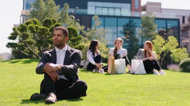 W idealny słoneczny dzień charyzmatyczny pracownik biurowy patrząc prosto w kamerę i pozując w garniturze siedzi na trawie w ogrodzie centrum biznesowego — Wideo stockowe