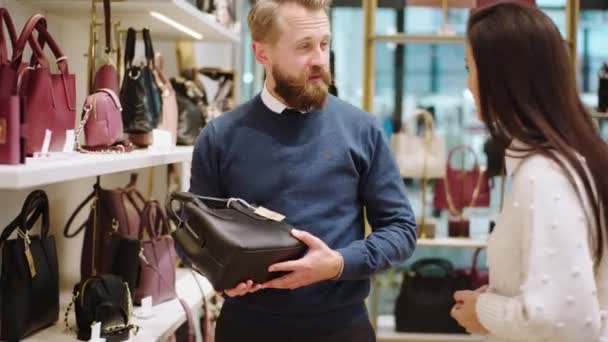 Hombre guapo cliente tratar de elegir una bolsa de la tienda de accesorios, mientras que una hermosa vendedora tratar de explicar algo de información sobre el producto. Disparo en ARRI Alexa Mini. — Vídeo de stock