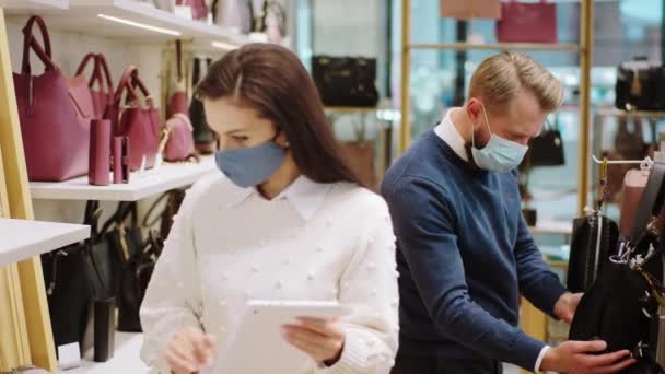 In the accessories store good looking saleswoman with protective mask using digital tablet to check the products from online while other supervisor check the quality of bags both wearing protective — 图库视频影像